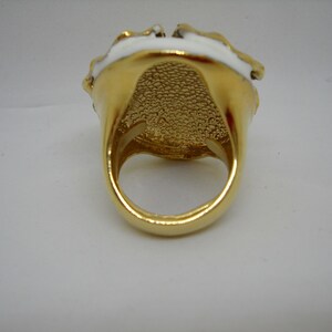 May include: A gold ring with a white enamel design. The ring has a textured band and a large, round face.