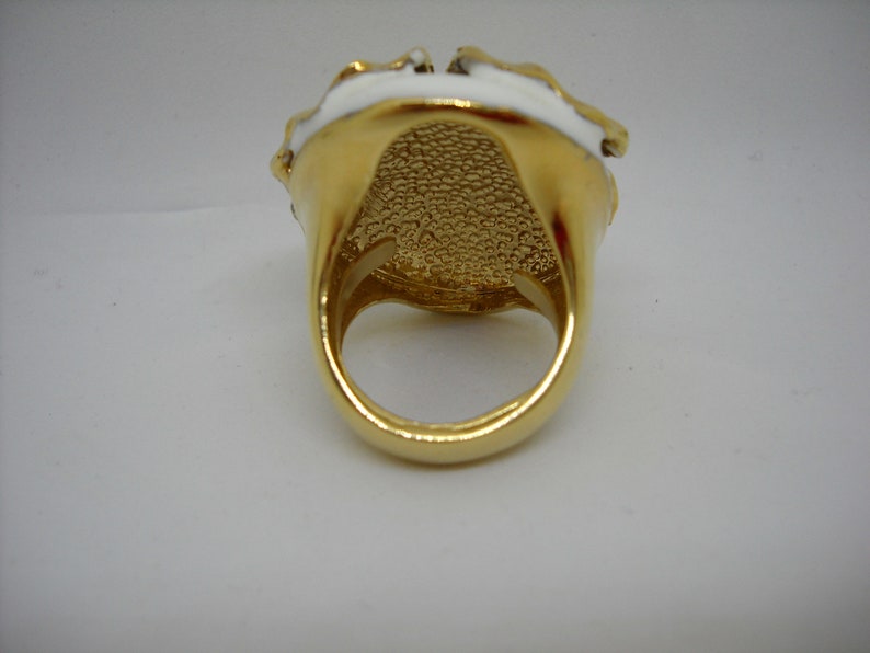 May include: A gold ring with a white enamel design. The ring has a textured band and a large, round face.