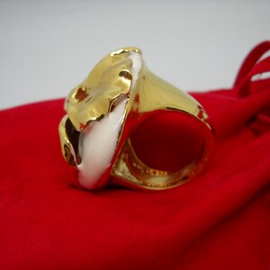 May include: A gold ring with a white ceramic design. The ring is on a red velvet surface.