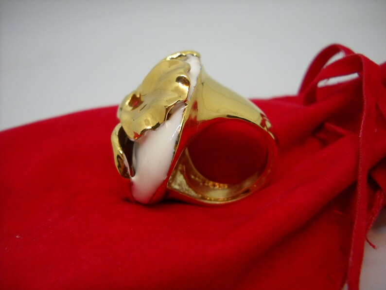 May include: A gold ring with a white ceramic design. The ring is on a red velvet surface.