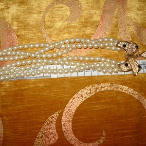 Op de afbeelding: Een meerstrengs parelarmband met goudkleurige sluitingen. De armband is ongeveer 15 cm lang. De sluiting heeft een decoratief strikdesign met zwarte accenten.