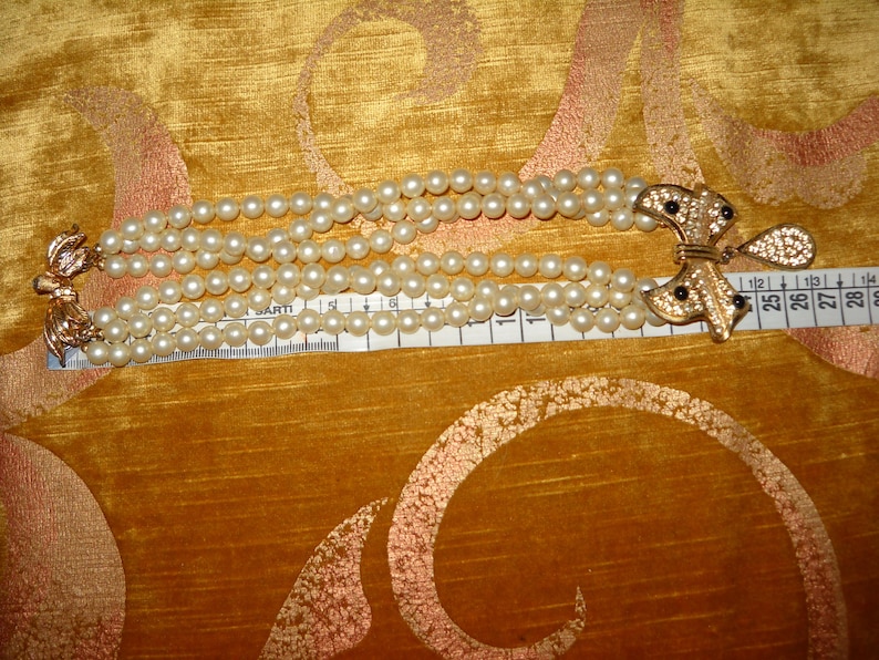 Op de afbeelding: Een meerstrengs parelarmband met goudkleurige sluitingen. De armband is ongeveer 15 cm lang. De sluiting heeft een decoratief strikdesign met zwarte accenten.