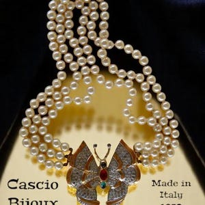 Puede incluir: Un colgante de mariposa dorado con piedras preciosas de colores, unido a un collar de perlas de múltiples hilos. El colgante tiene el texto "Cascio Bijoux" y "Made in Italy 1970" en la parte inferior.