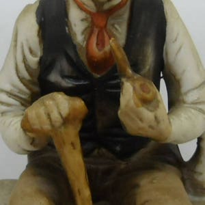 May include: A porcelain figurine of a man wearing a vest and holding a walking stick and a pipe. He is sitting on a base and has a detailed facial expression.