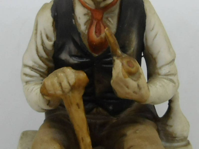 May include: A porcelain figurine of a man wearing a vest and holding a walking stick and a pipe. He is sitting on a base and has a detailed facial expression.