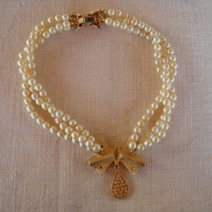 Op de afbeelding: Een parelketting met drie strengen, een goudkleurige strik-hanger en een druppelvormige bedel. De ketting heeft een gouden sluiting. De parels zijn cr&egrave;mewit. Neutrale achtergrond.