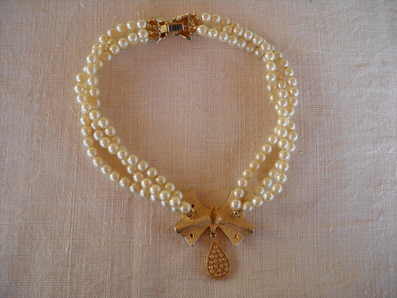 Op de afbeelding: Een parelketting met drie strengen, een goudkleurige strik-hanger en een druppelvormige bedel. De ketting heeft een gouden sluiting. De parels zijn cr&egrave;mewit. Neutrale achtergrond.