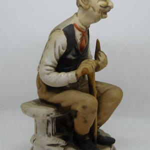 May include: A porcelain figurine of a man with a white beard and mustache, wearing a brown vest and sitting on a white bench. He is holding a walking stick in his right hand.