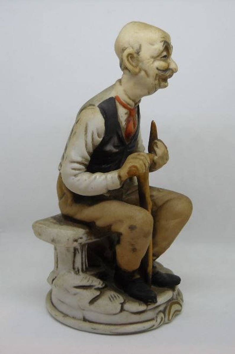 May include: A porcelain figurine of a man with a white beard and mustache, wearing a brown vest and sitting on a white bench. He is holding a walking stick in his right hand.