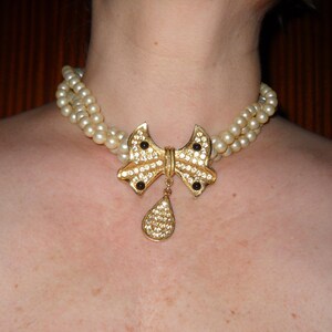 Op de afbeelding: Een vintage parelketting met een goudkleurige strik-hanger. De strik is versierd met heldere en zwarte strass-steentjes, en een druppelvormige hanger hangt eronder. De ketting heeft twee strengen imitatieparels.