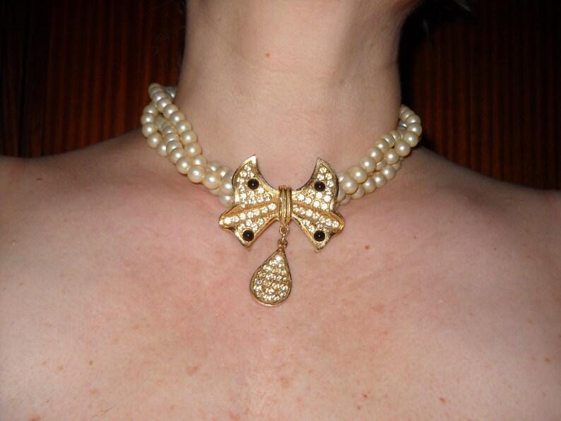 Op de afbeelding: Een vintage parelketting met een goudkleurige strik-hanger. De strik is versierd met heldere en zwarte strass-steentjes, en een druppelvormige hanger hangt eronder. De ketting heeft twee strengen imitatieparels.