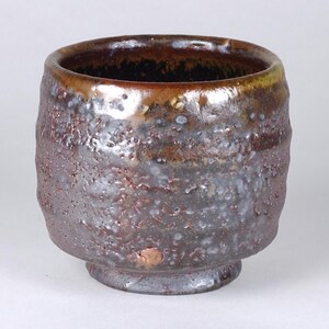 May include: A handcrafted ceramic cup with a textured, rough surface. The cup features a dark brown exterior with lighter brown and white accents. The rim is a darker shade of brown. The cup has a small, round base.