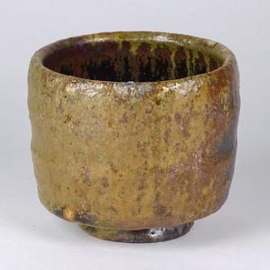 May include: A handcrafted, rustic ceramic bowl with a textured, earthy appearance. The bowl features a rounded shape with a slightly flared rim and a small, stable base. The exterior is primarily a mottled, muted brown with hints of green and gold.