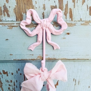 May include: A pink metal hook shaped like a bow, mounted on a light blue wooden surface. A pink fabric bow is hanging from the hook.