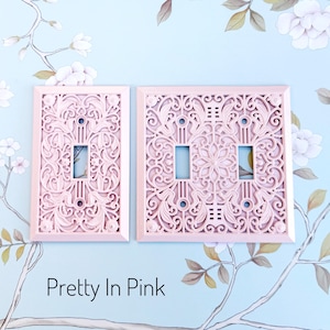 K&ouml;nnte beinhalten: Zwei rosa dekorative Lichtschalterabdeckungen mit aufwendigen Blumen- und Rankenmustern. Die Abdeckungen sind rechteckig mit Aussparungen f&uuml;r die Schalter. Der Text "Pretty In Pink" steht unten.