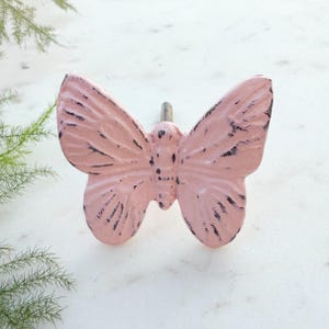 May include: A pink butterfly-shaped cabinet knob with a distressed finish. The knob is made of metal and has a screw for attaching it to a cabinet door.