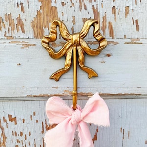 May include: Gold metal hook shaped like a bow with a pink fabric bow hanging from the hook.