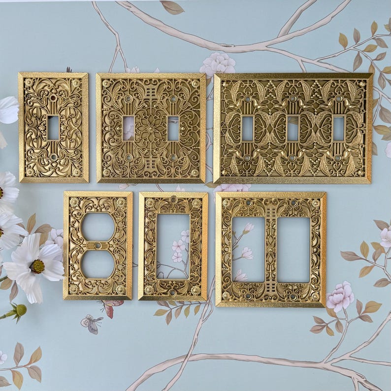 K&ouml;nnte beinhalten: Eine Sammlung verzierter, goldfarbener Lichtschalterplatten. Die Platten weisen aufwendige Blumen- und Rankenmuster auf. Verschiedene Konfigurationen umfassen Einzel-, Doppel- und Dreifachschalterplatten sowie eine Steckdosenplatte.