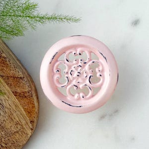 May include: A round, pink, metal cabinet knob with a distressed finish. The knob has a decorative, openwork design in the center.