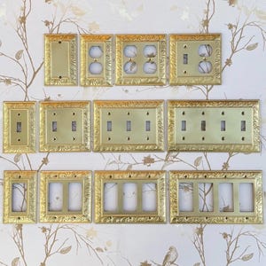 May include: A collection of decorative brass switch plates and outlet covers. The plates feature ornate, embossed frames in various configurations, including single switch, double switch, and multiple outlet designs. The plates are displayed against a wallpaper background with a floral pattern.