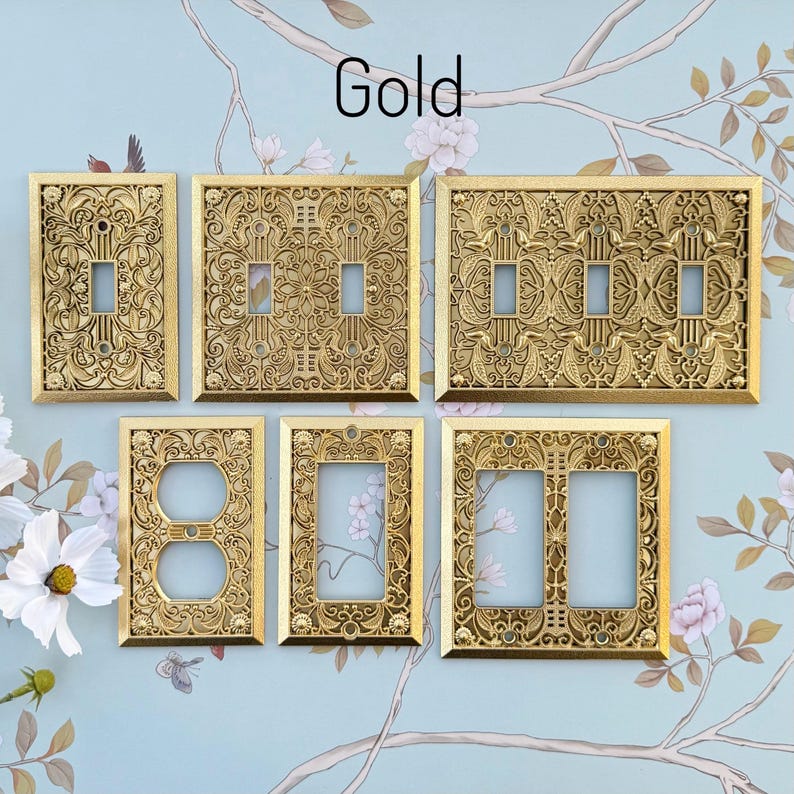 K&ouml;nnte beinhalten: Eine Sammlung verzierter, goldfarbener Lichtschalter- und Steckdosenabdeckungen. Die Abdeckungen weisen aufwendige Blumen- und Rankenmuster auf. Das Wort "Gold" ist in schwarzer Schrift oben geschrieben. Sie sind auf hellblauem Hintergrund mit floralen Mustern angeordnet.