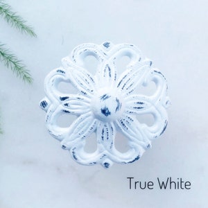 May include: White metal cabinet knob with a distressed finish and a floral design. The knob is round and has a raised center. The text "True White" is visible below the knob.