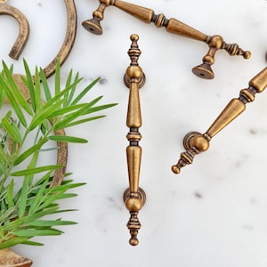 May include: An overhead shot showcases several antique brass cabinet pulls and handles. The hardware features ornate detailing and a warm, aged finish. The arrangement includes a variety of sizes and styles, suggesting a collection for home decor or restoration projects.