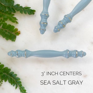 May include: Three blue cabinet knobs with gold accents. The knobs are 3 inches apart. The knobs are painted in a color called "Sea Salt Gray".