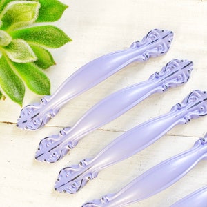 May include: Five lavender-colored cabinet knobs with ornate, black-painted details. The knobs have a slightly distressed finish and are arranged in a row.