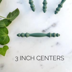 May include: A green metal cabinet pull with a 3 inch center to center measurement. The pull has a decorative design on each end.