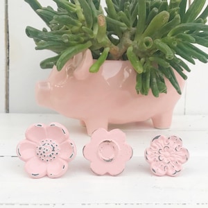 May include: Three pink flower-shaped drawer pulls with distressed white paint. The pulls are arranged in a row on a white surface.