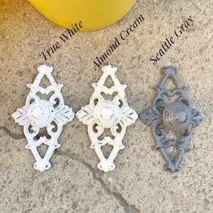 May include: Three decorative cabinet knobs in different colors: True White, Almond Cream, and Seattle Gray. Each knob is a diamond shape with a round center and intricate details.