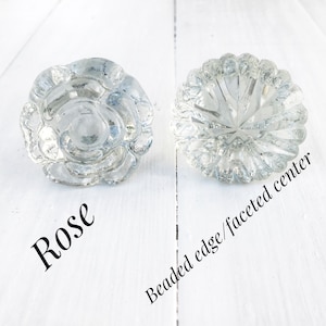 May include: Two clear glass cabinet knobs. One is a rose design and the other has a beaded edge and faceted center.