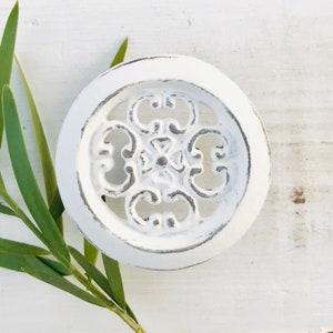 May include: White round decorative knob with a detailed, ornate, floral design. The knob has a distressed, aged look.