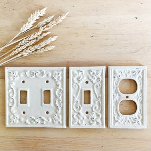 May include: Three white ceramic light switch and outlet covers with an ornate design. The covers have a distressed white finish and are in a farmhouse style.
