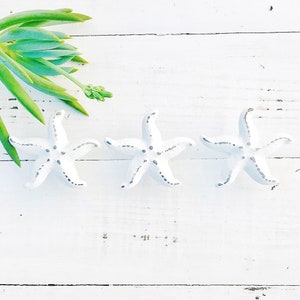 May include: Three white ceramic starfish with gray accents on a white wooden background. The starfish are arranged in a row.
