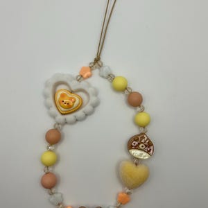 May include: A beaded phone charm featuring a variety of pastel-colored beads, including yellow, peach, and clear beads. The charm includes a white heart-shaped embellishment with a bear design, a brown bear charm, and a yellow heart.