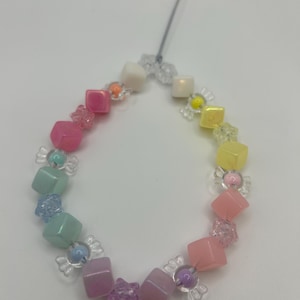 Puede incluir: Un collar de cuentas de colores arcoíris con cuentas de forma cuadrada y cuentas transparentes en forma de caramelo. El collar está hecho con cuentas de color rosa pastel, azul claro, amarillo y blanco.