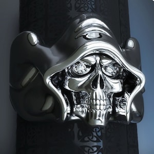 May include: A silver skull ring with a hooded figure design. The skull has two large, clear gemstones for eyes. The ring is made of metal and has a detailed design.