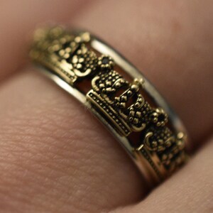 May include: A silver and gold ring featuring a repeating crown design. The ring is worn on a finger.
