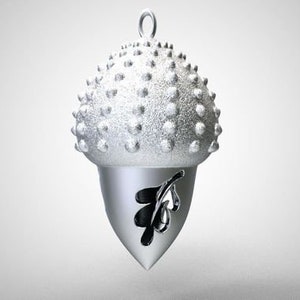 Puede incluir: Un amuleto de plata en forma de bellota con una superficie texturizada detallada y un diseño de hoja recortada.