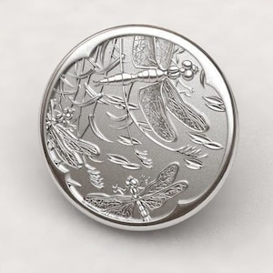 Könnte beinhalten: Eine runde silberne Schmuckdose mit einem geprägten Design aus drei Libellen. Die Libellen befinden sich in einer natürlichen Umgebung mit Blättern und Stielen.