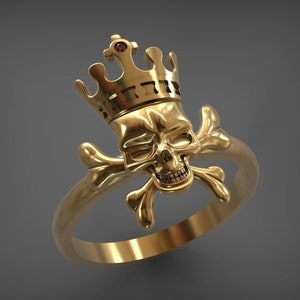 Op de afbeelding: Een goudkleurige ring met een schedel met een kroon en gekruiste botten. De kroon heeft een kleine rode edelsteen. De ring staat tegen een grijze achtergrond. Een opvallend sieraad.