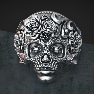 May include: Silver ring with a detailed skull design featuring floral accents and intricate patterns. The skull has a feminine appearance with delicate features and a floral crown.