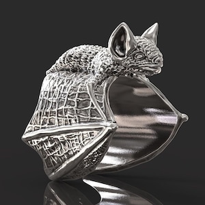 以下が含まれることがあります： 銀色のコウモリの形をしたリング。コウモリは翼を広げた状態で描かれており、リングの構造を形成しています。コウモリの体はテクスチャ加工された表面を持っています。リングは暗い背景に配置されています。