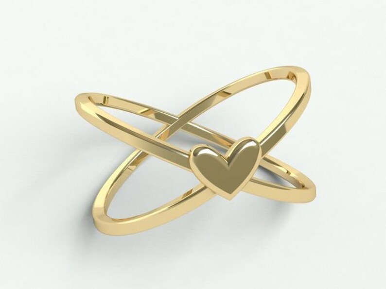 K&ouml;nnte beinhalten: Ein goldfarbener Ring mit einem sich kreuzenden Design, das zwei B&auml;nder und einen herzf&ouml;rmigen Akzent aufweist. Der Ring hat eine polierte Oberfl&auml;che und ist vor einem wei&szlig;en Hintergrund platziert. Das Herz befindet sich in der Mitte der sich kreuzenden B&auml;nder.