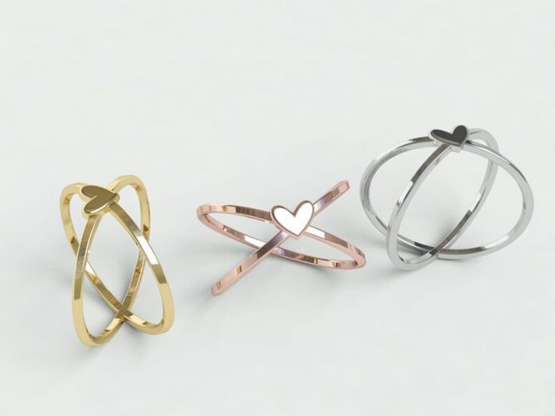 K&ouml;nnte beinhalten: Drei Ringe mit Herzdesign. Ein Ring ist goldfarben, einer ros&eacute;goldfarben mit wei&szlig;em Herz und der dritte silberfarben mit schwarzem Herz. Die Ringe haben ein sich kreuzendes Banddesign.