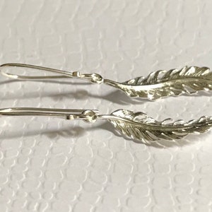 Może przedstawiać: Para srebrnych kolczyków w kształcie piór. Każdy kolczyk ma szczegółowy wzór pióra z zapięciem na haczyk. Kolczyki są prezentowane na białej, teksturowanej powierzchni, ukazując ich delikatne rzemiosło.