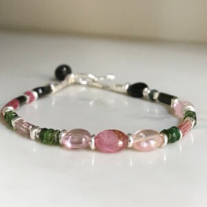 May include: A delicate bracelet featuring pink, green, and black gemstones, with silver accents. The bracelet has a simple clasp closure.