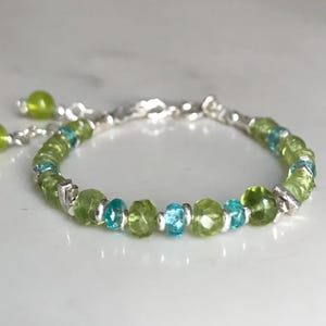 May include: A delicate bracelet featuring a combination of green and blue gemstones. The bracelet is made with small, faceted peridot beads and interspersed with turquoise-colored beads. The silver chain has a textured design and includes two dangling green beads.